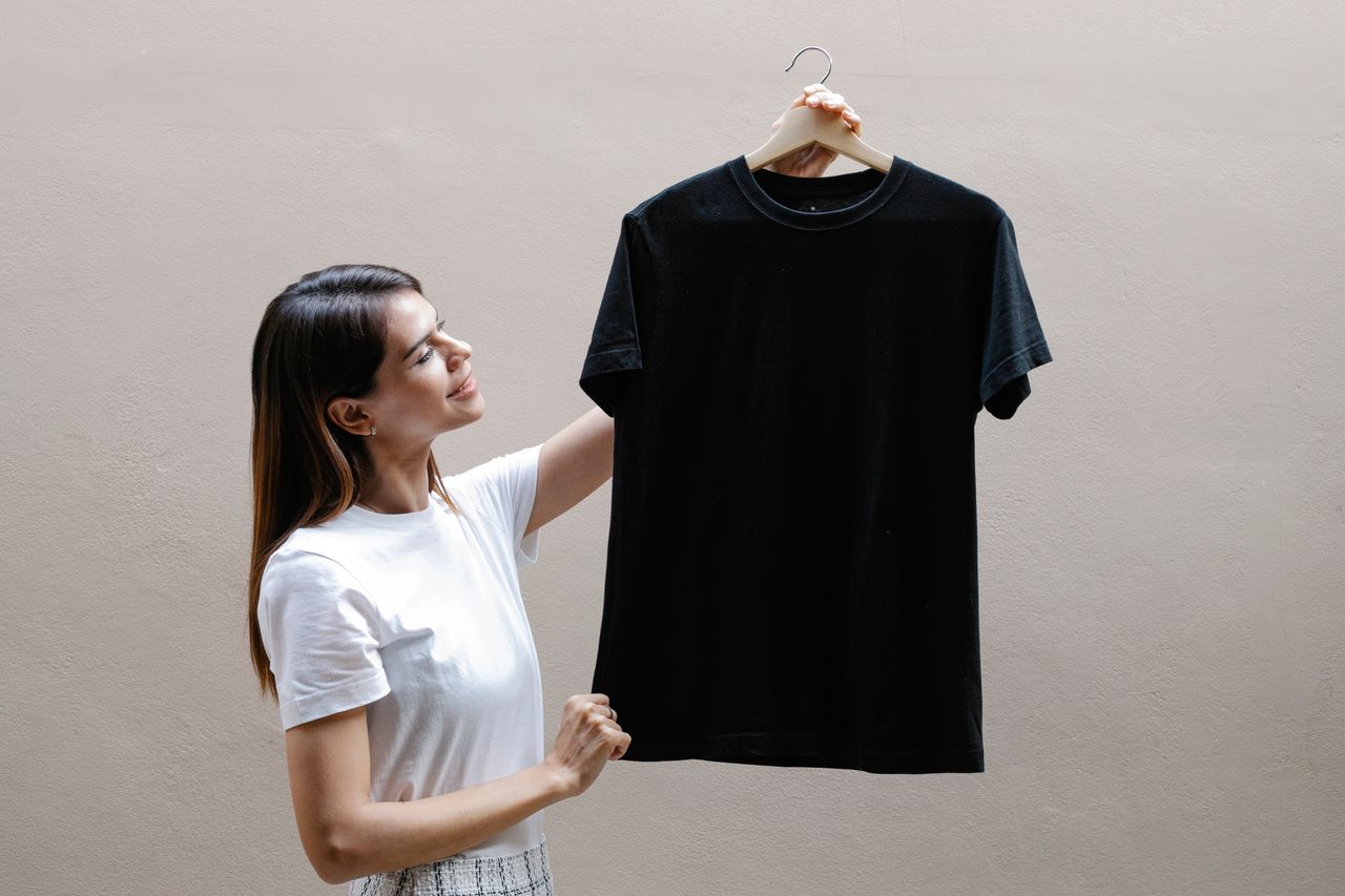 Side view of self assured young woman with long dark hair in stylish outfit smiling while holding hanger with black t shirt against gray wall